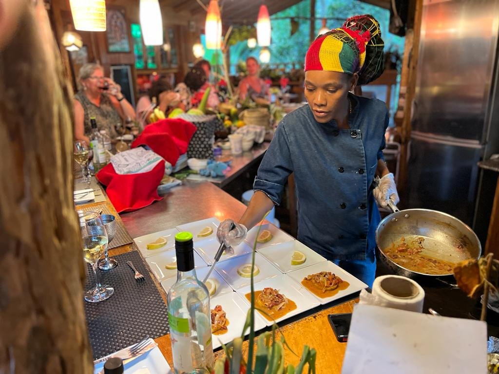 Chef Alecia Swainbank in a Rasta-colored headwrap and denim chef coat, plating pepper shrimp during a live dinner service.