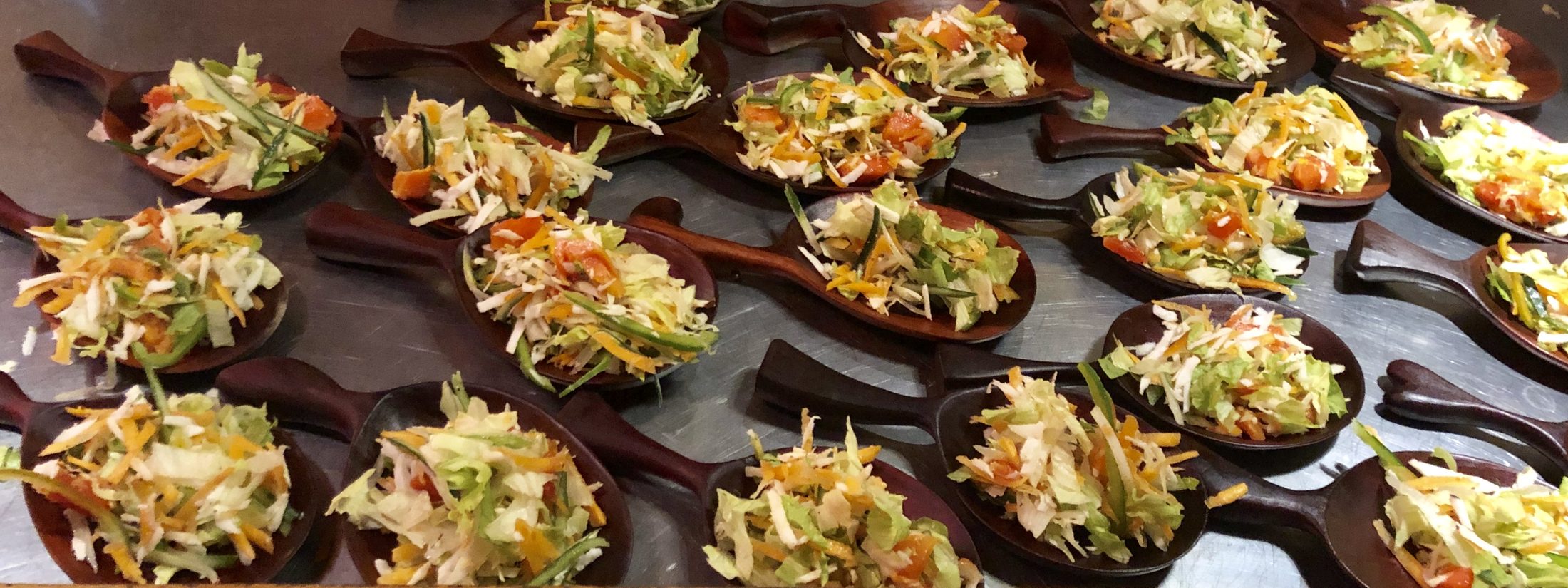 A wide arrangement of hand-carved wooden spoons filled with chopped garden salad — julienned carrot, cucumber ribbons, tomato, and greens.