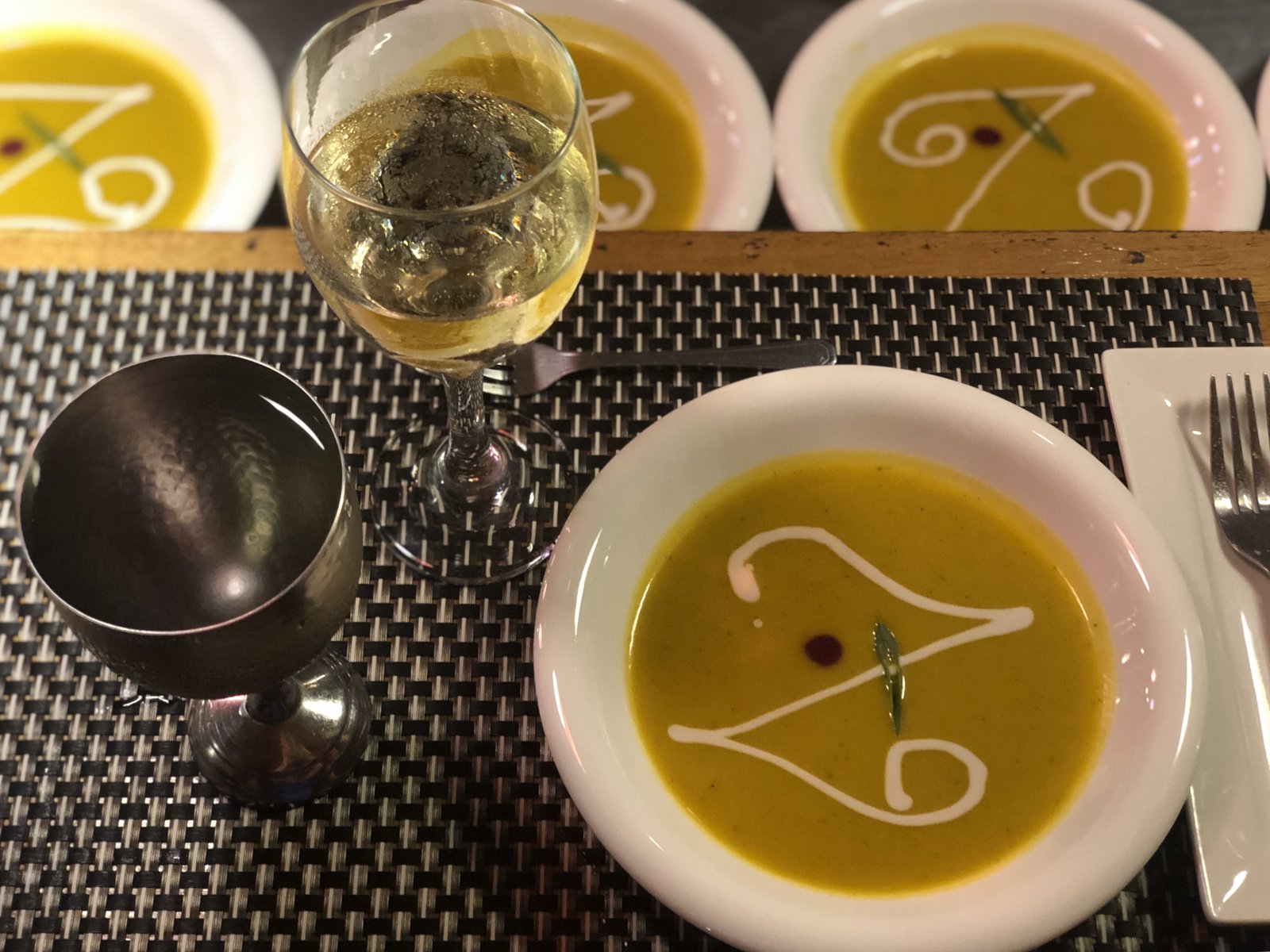 Four plates of golden pumpkin soup finished with a signature Z-shaped cream swirl, on a woven placemat with a hammered goblet.