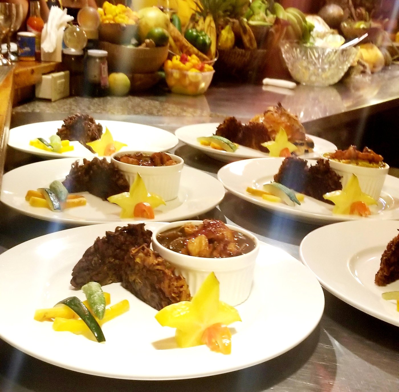 A line of white plates dressed with star-fruit garnish and pickled vegetable sides, with a wall of fresh tropical fruit behind.
